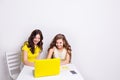 Two girls sit behind a laptop in yellow case. There is a smartphone on the table charging via yellow cable. Both girls