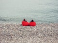 Two girls sit in red armchair bags on the pebble sea shore on a cool spring day