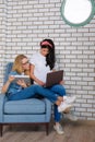 Two girls sitting on a chair in bright office and  working with different gadgets. Brunette and blonde check work email on the tab Royalty Free Stock Photo