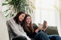 Two girls sitting at sofa, watching something funny at tablet, smiling, laughing