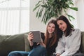 Two girls sitting at sofa, watching something funny at tablet, smiling, laughing