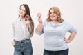 Two girls, thin and fat on a white background, are holding pink glazed donuts in their hands.