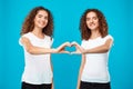 Two girls twins showing heart with hands over blue background.