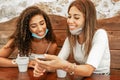 Two girls wearing protection mask sitting at bar table looking smartphone with coffee cups. New normal human habits: having fun Royalty Free Stock Photo