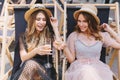 Two glad girls in the same straw hats celebrating holiday with champagne chilling in recliners. Outdoor portrait of