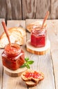 Two glass jars of strawberry and apple jam with spoons and sliced bread on wooden table