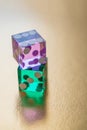 Two glass multi-colored dice on a gold background in sunlight. The pink die with a score of six lies on the green die.