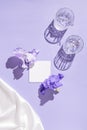 Two glasse with water and card note with flowers on pastel lilac background with white silk cloth. Summer refreshment concept