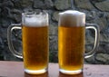 Two glasses of cold draft beer with lush foam on a wooden desk with a stone wall background