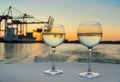 Two glasses of cool white wine on white tablecloth with harbor structures and cargo ship in blurred background