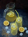 Two glasses and jug of cold homemade lemonade with lemon slices, ice cubes, brown sugar, yellow straws
