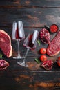 Two glasses of red wine near raw beef steaks with seasonings over rustic dark wood background, top view