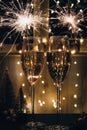 Two glasses of sparkling wine champagne with sparklers. Dark background with yellow light bokeh. Christmas tree toys on