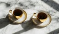 Two golden teacups filled with dark beverage sit on saucers on marble surface. Sunlight creates shadows, reflections on Royalty Free Stock Photo