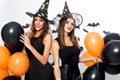 Two gorgious women in black dresses and witch hats hold black and orange balloons. Halloween