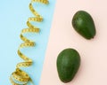 Two green avocado fruits and a twisted measuring yellow tape on a beige-blue background