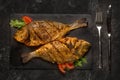 Two grilled dorado fish on dark black granite stone board on black table.