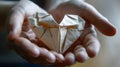 Two hands clasping a small intricate and perfectly folded origami heart with delicate paper fibers visible in the