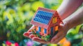 two hands cradling a small model house adorned with solar panels on the roof, against a backdrop of blurred greenery