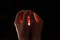 Two hands folded squeeze the light inside, reddish light inside the hands, black background.