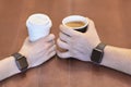 Two hands, male and female, both with equal electronic wrist watches, holding cups of coffee, white and black, on the wooden table Royalty Free Stock Photo