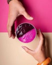 Two hands, one with an orange bangle, hold a plastic cup containing a halfeaten dragon fruit dessert, with one side purple and the Royalty Free Stock Photo