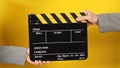 Two hands send and hold a Black clapper board on yellow background. Arms wear a grey suit