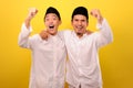 Two handsome happy Asian Muslim men hugging and looking at camera with satisfied amazed face while showing winning gesture Royalty Free Stock Photo