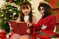 Two happy beautiful little girl reading a storybook together in a cozy living room on Christmas eve