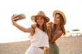 Two happy blonde caucasian sisters having fun on the beach during summer, taking selfie with mobile phone, smiling and looking to Royalty Free Stock Photo
