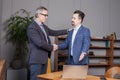 Two happy business partners in blue suits smiling and making handshake in office. Businessman pointing on the laptop Royalty Free Stock Photo