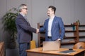 Two happy cherrful businessman in blue suits smiling and making handshake in office by the laptop, business succes concept