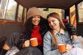 Two happy multiracial girlfriends on car trunk in autumn forest, embracing and drinking hot tea Royalty Free Stock Photo