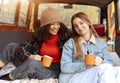Two happy multiracial girlfriends on car trunk in fall forest, embracing and drinking hot tea during adventure trip in autumn Royalty Free Stock Photo