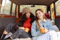 Two happy multiracial girlfriends sitting on car trunk and drinking tea during adventure road trip in nature Royalty Free Stock Photo