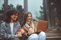 Two happy multiracial teen girls looking at laptop and laughing while drinking coffee in autumn park Royalty Free Stock Photo