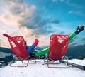 Two happy skiers have a relax time in red chairs on top of mountain