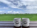 Two happy smiling cups of coffee or tea on the wooden table. With bright blue summer sky and clouds over the green field. Royalty Free Stock Photo