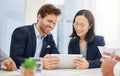 Two happy young diverse colleagues discussing plans and ideas together on a digital tablet device during a meeting in an Royalty Free Stock Photo