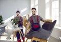 Two happy young moving company workers carrying chairs, armchairs and house plants