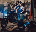 Two brunette women in blue overalls posing next to a custom bobber in authentic workshop garage Royalty Free Stock Photo