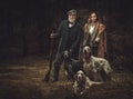 Two hunters with dogs and shotguns in a traditional shooting clothing, posing on a dark forest background. Royalty Free Stock Photo