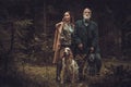 Two hunters with dogs and shotguns in a traditional shooting clothing, posing on a dark forest background. Royalty Free Stock Photo