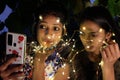 Indian female friends holding illuminated string lights and taking photos in the evening