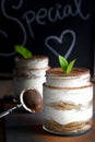 Two jars filled with tiramisu with cocoa powder on top and mint leaves as decoration on a chalkboard background. Dark food concept