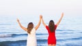 Two joyful girls girlfriends standing on the beach looking at the sea horizon with hands up