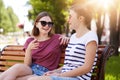 Two joyful girls relax together in local park sitting on new wooden bench, make jokes, gossip, remember old funny moments with