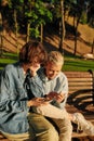 Two joyful women holding their smartphone, sitting on the bench in the park. Lesbian couple using their phones while Royalty Free Stock Photo