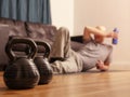 Two kettle bells in focus. Bald man laying on the floor of living room with a blue bottle of water in his hand out of focus, Royalty Free Stock Photo