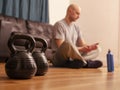 Two kettle bells in focus. Bald man sitting on a floor of living room checking his pulse out of focus, Concept Work out at home,